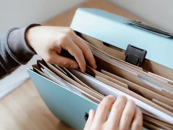 Pessoa organizando documentos em uma pasta sanfonada azul, com papéis separados por divisórias sobre uma mesa.
