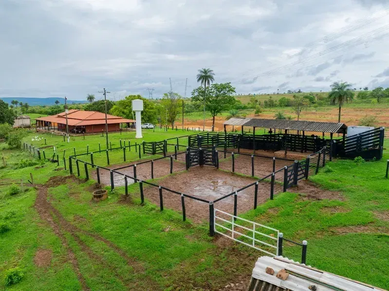 Vista aérea de uma propriedade rural com curral cercado por madeira, área de manejo de gado, pasto verde ao redor, casa de fazenda com telhado vermelho ao fundo e paisagem campestre com árvores e colinas sob céu nublado.